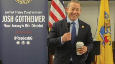 Rep. Josh Gottheimer stands in front of banners with his name on it, holding a Starbucks hot drink in one hand and a microphone in the other.