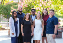 A group of six students stand together smiling in front of a green background.