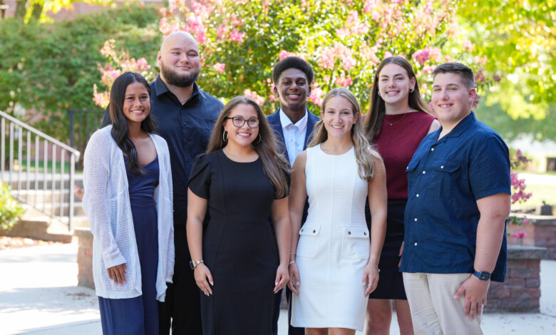 A group of six students stand together smiling in front of a green background.